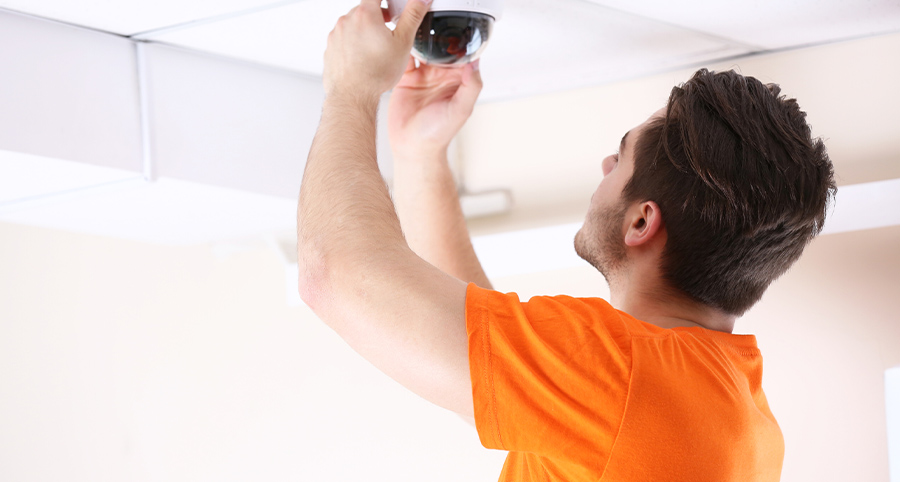 A security tech man in an orange shirt installing a ceiling camera in a business setting.